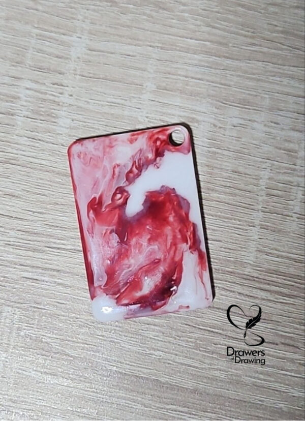 Dettaglio di una targhetta in resina con sfumature fluide rosse e bianche appoggiata su un piano in legno con logo Drawers of Drawing
